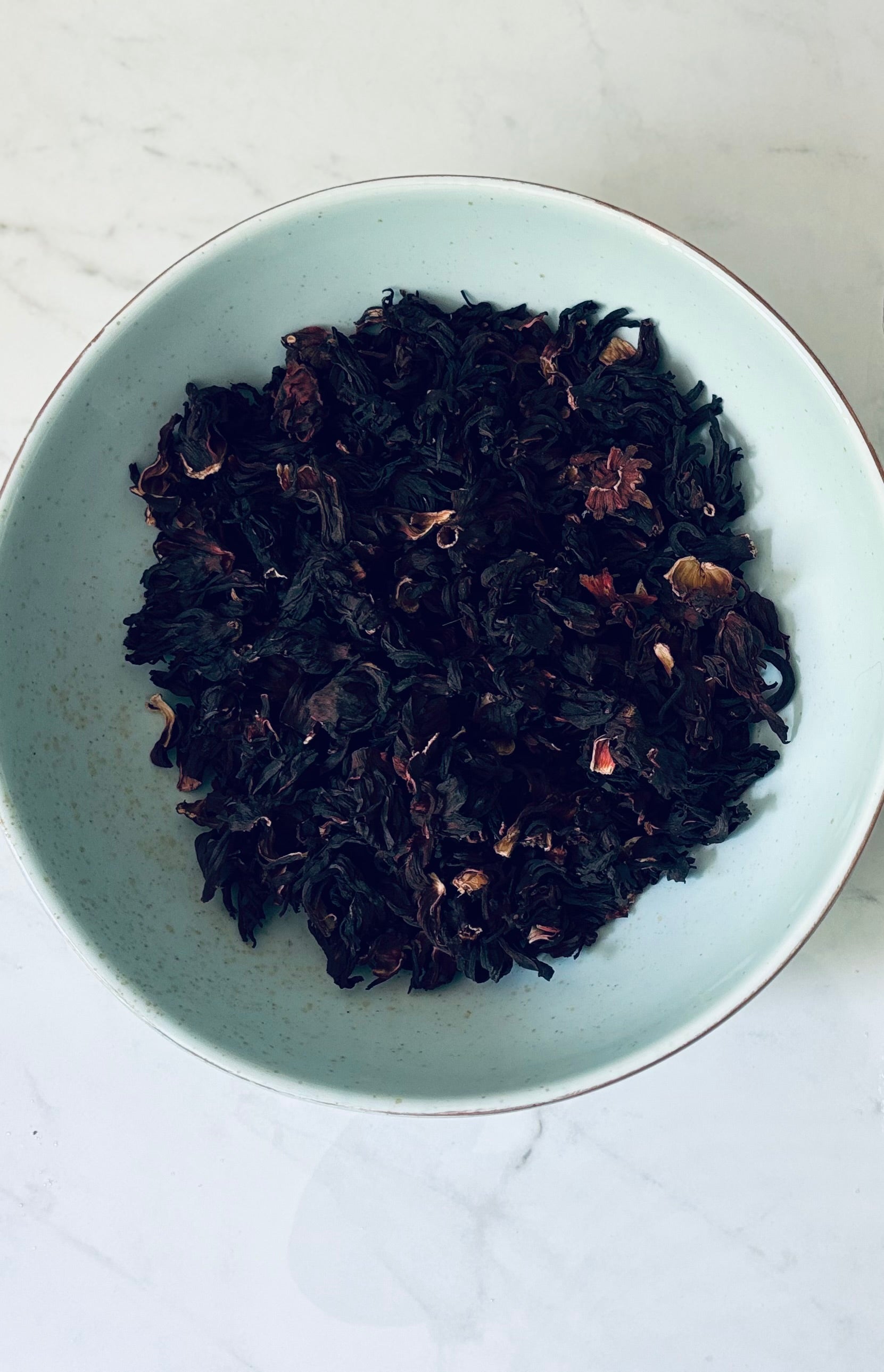 Dried hibiscus buds in a light blue bowl on a white marble surface. Image is placed above a statement about the product ingredients. 