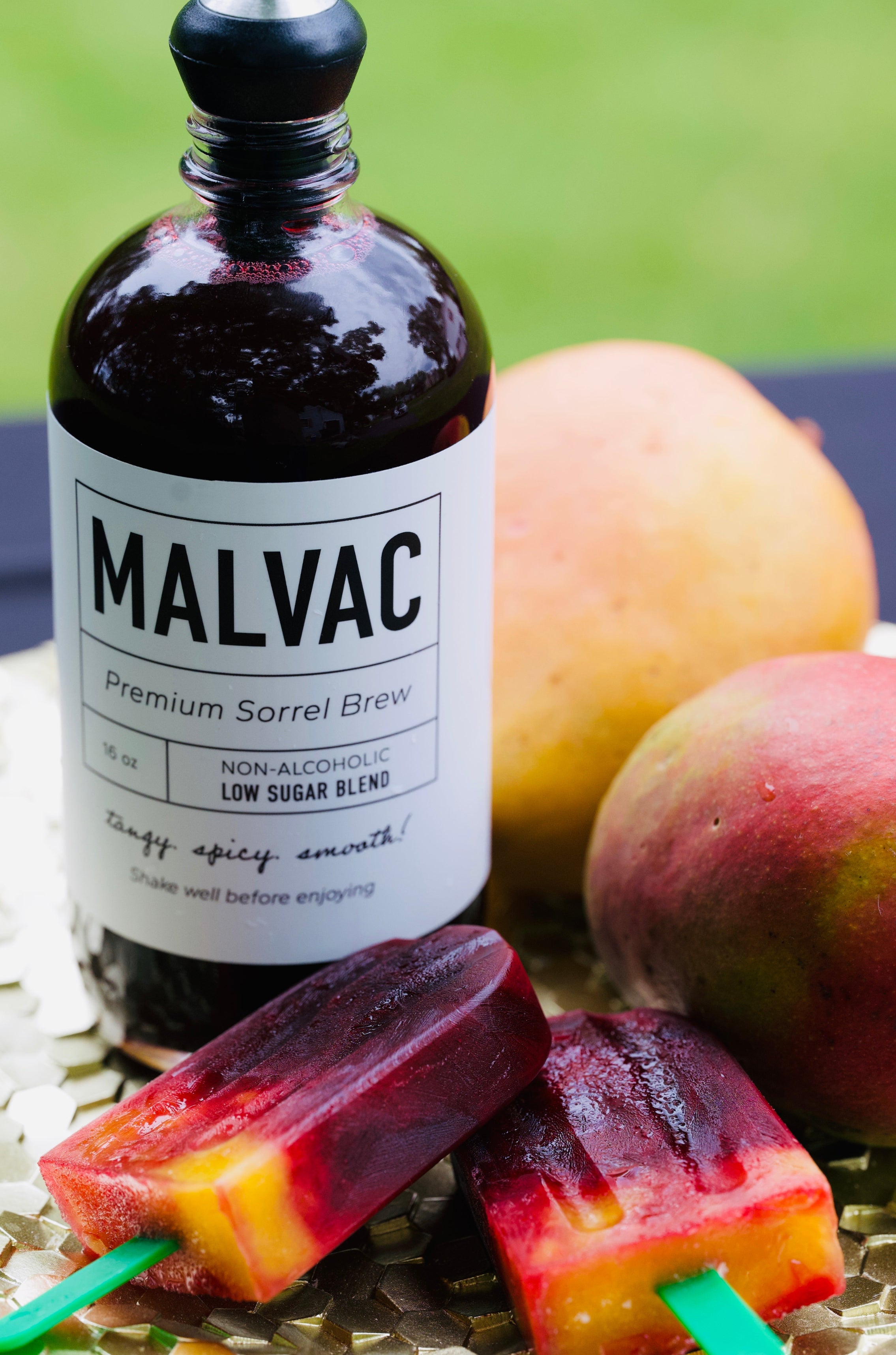 Bottle of MALVAC Premium Sorrel Brew with two mangoes and two popsicles on a table outdoors.
