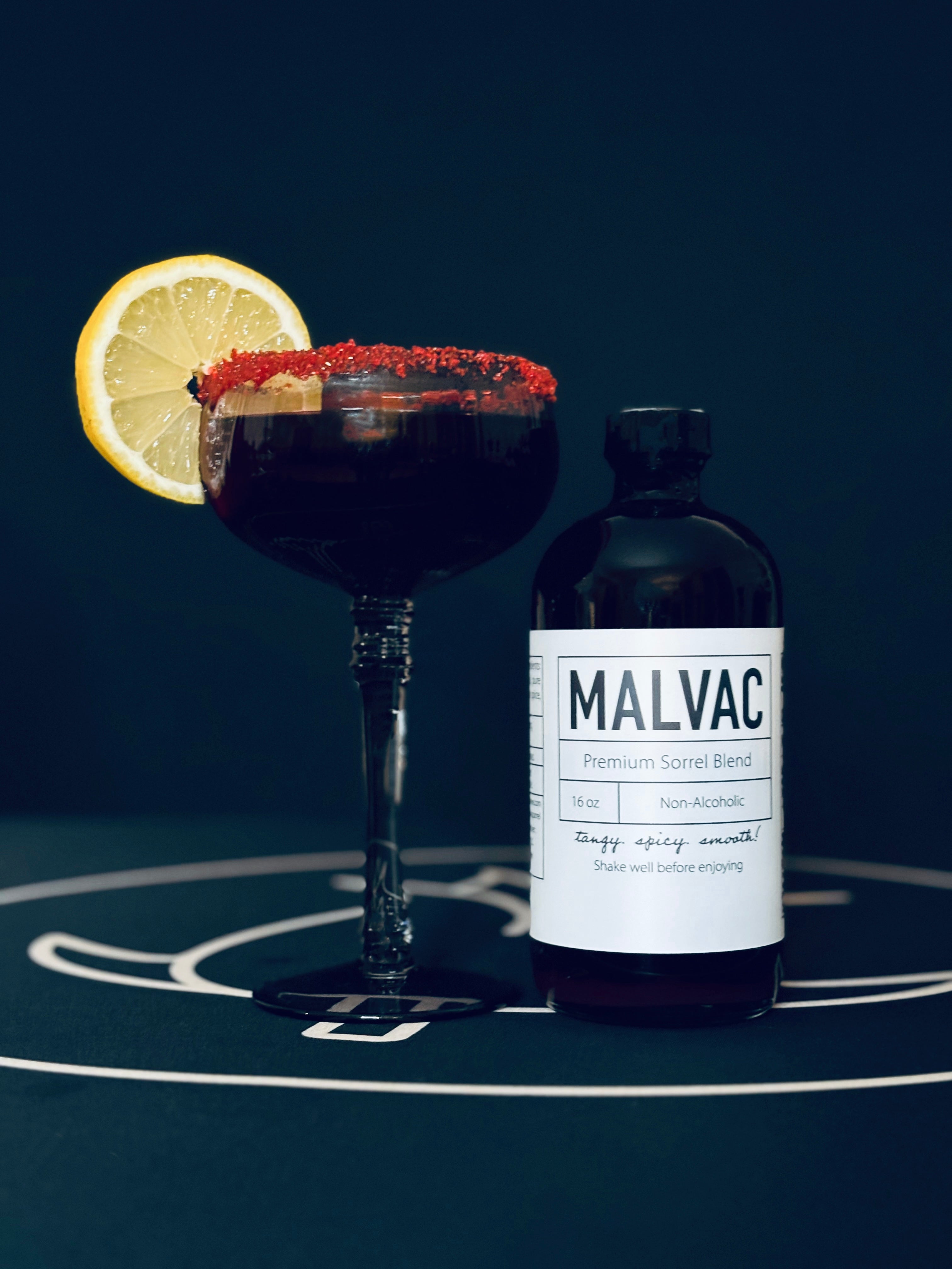 Bottle of MALVAC Premium Sorrel Brew with a cocktail with a lemon wedge and red sugar rim on a dark background.